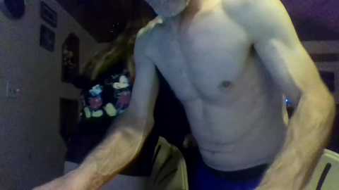 Video of a shirtless man with a thin, muscular physique, wearing blue shorts. He stands indoors, with a flat-screen TV displaying a colorful cartoon in the background.