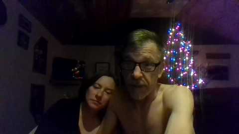 A dimly-lit video shows a shirtless, middle-aged man with glasses and a woman with shoulder-length hair, both looking tired, in a cluttered living room with a Christmas tree.