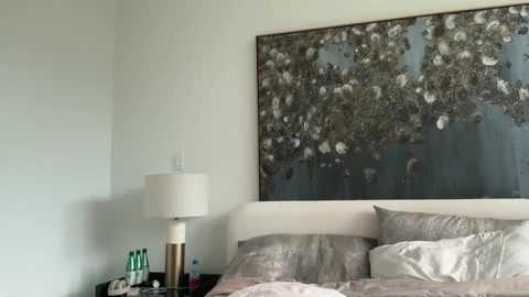Video of a serene bedroom featuring a large, abstract tree painting, light beige walls, a white bedside lamp, and a neatly made bed with silver and white bedding.