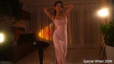 Video of a slender woman with long dark hair in a tight, pink, floor-length evening gown, posing with hands behind her head, in a dimly lit, modern living room with warm lighting, green plants, and decorative elements.