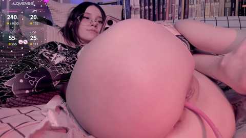 A video of a young woman with glasses and a black floral dress, lying on a bed with a pink thong visible, surrounded by books and a video game console.