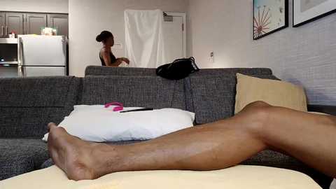 Video of a man lying on a gray sectional couch, legs extended, in a modern living room with white walls, kitchen visible in the background.