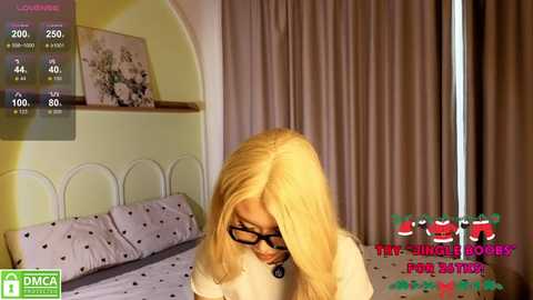 Video of a blonde woman with glasses, wearing a white sweater, sitting on a bed with polka-dotted sheets. Background features a wall with floral artwork and beige curtains.