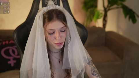 Media: Video of a fair-skinned woman with long brown hair in a white veil, sitting in a black gaming chair, with a tattooed arm and a green plant in the background.