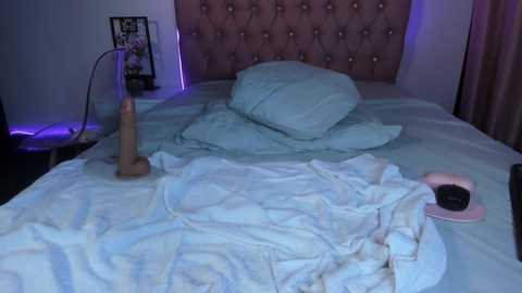 Video of a messy bedroom with a tufted pink headboard, disheveled white bedding, a sex toy, and a pair of headphones on a nightstand, bathed in blue and purple lighting.