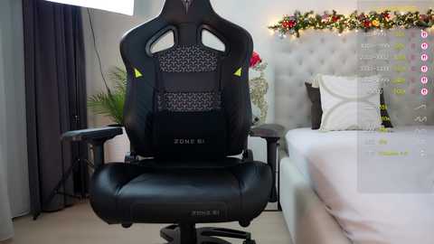 Video of a modern gaming chair with a black mesh back, situated in a cozy, well-lit bedroom with a tufted headboard, floral garland, and white bedding.