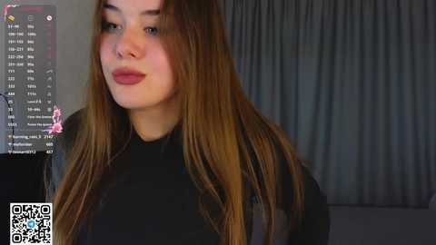 A video of a young woman with long, straight brown hair, wearing a black top, smiling slightly, standing in front of a computer monitor displaying a chat interface.