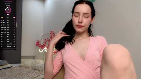 Video of a fair-skinned woman with long black hair, wearing a pink V-neck top, sitting in a bedroom with pink flowers and a digital clock in the background.