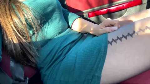 Video of a woman in a teal dress with long brown hair, sitting in a car. She has a tattoo of a heartbeat on her thigh. Red car seat in the background.