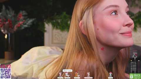 Video of a young woman with long, straight blonde hair, fair skin, and a slight smile, sitting indoors. Background features a blurred green plant and a wooden table.