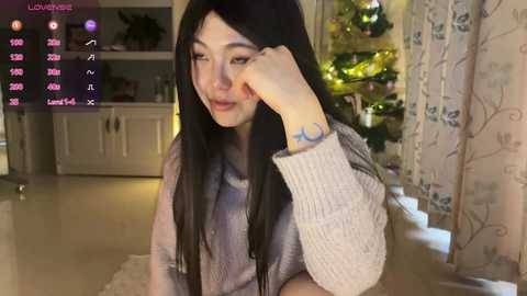 Video of an Asian woman with long black hair, fair skin, and a tattoo on her wrist, wearing a beige sweater, sitting indoors near a decorated Christmas tree.