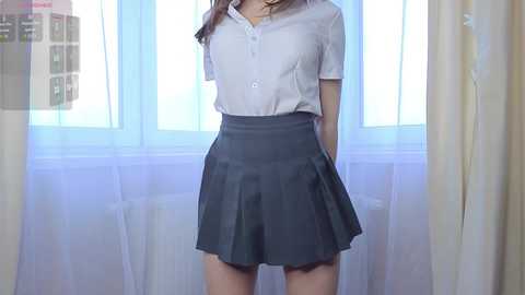 Media: Video of an Asian woman in a white blouse and grey pleated skirt, standing in front of a window with white curtains.