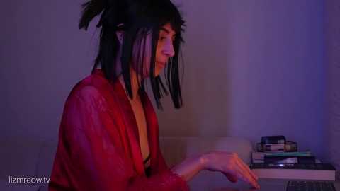 A video of a person with long, black, frayed hair, wearing a red robe, sitting in a dimly lit room, holding a book.