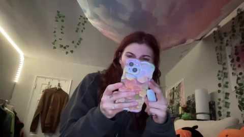 Video of a young woman with long, straight red hair, wearing a dark hoodie, taking a selfie with a pink camera phone in a dimly lit room adorned with green ivy wallpaper, fairy lights, and a Halloween-themed plush toy.