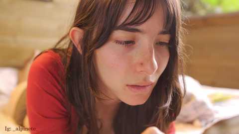 Video of a young woman with long brown hair, wearing a red top, looking contemplative, in a warm, blurred indoor setting.