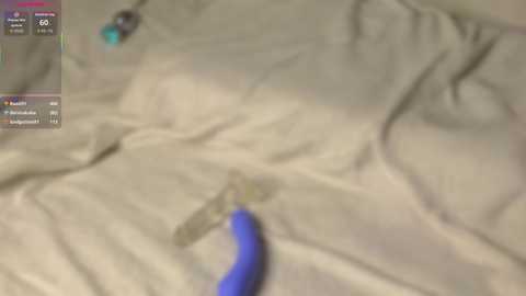 A blurry, low-resolution image showing a person's arm and hand holding a blue object on beige fabric, possibly a bed sheet, with a video game interface in the top left corner.