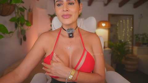 Video of a woman with short, dark hair, light brown skin, wearing a red bikini top, gold bracelet, and necklace, sitting in a cozy, dimly-lit room with potted plants and wooden furniture.