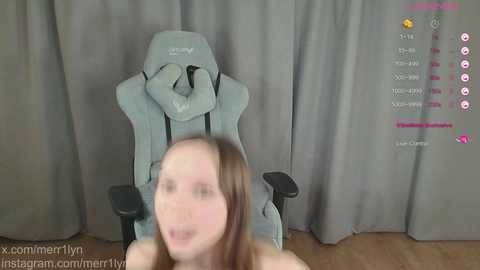 Video of a woman in a gaming chair, blurred face, streaming on twitch, grey curtains, user name \"xcommerlyn,\" wooden floor.