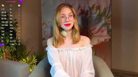 Video of a fair-skinned, blonde woman with glasses and red lipstick, wearing a white off-shoulder blouse, seated in a grey chair, in a room with floral wallpaper and a green plant.
