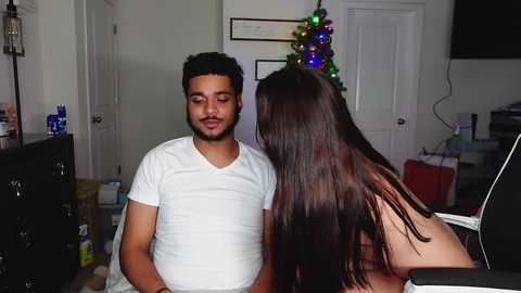 Media: Video of a young man with short black hair and medium skin tone wearing a white T-shirt, standing in a living room next to a woman with long brown hair, partially topless.