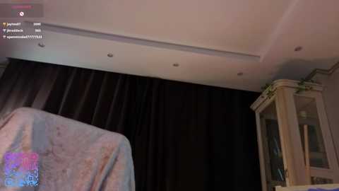 Media: A video of a dimly lit room with a white ceiling and black curtains. A man sits in a chair draped in a light blue towel. A glass cabinet is partially visible on the right.