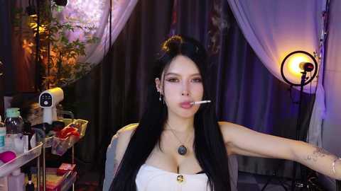 Video of a woman with long black hair and fair skin, wearing a strapless white top, sitting in a dimly lit room with a microphone, a light, and a desk with makeup items in the background.