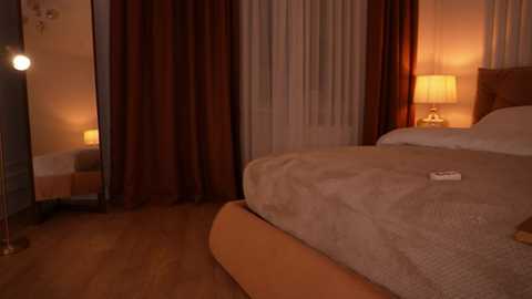 Video of a warmly lit bedroom with a beige bed, plush beige blanket, wooden floor, maroon and white curtains, bedside lamp, and a small white TV remote.