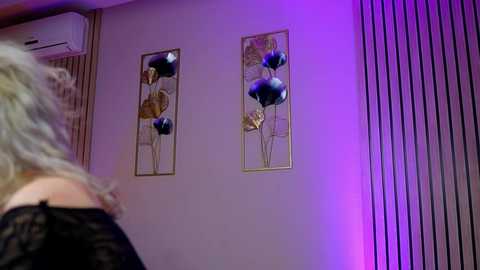 Media: Video of a woman with wavy blonde hair, standing in a room with two modern, gold-framed abstract flower artworks and purple accent lighting, focusing on her back.