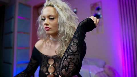 Video of a platinum blonde woman with wavy hair in a black, sheer lace bodysuit, posing seductively, surrounded by purple and blue lighting in a dimly lit room.