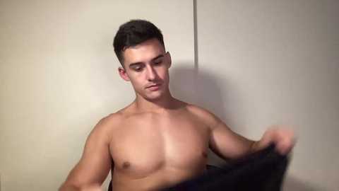 Video of a shirtless, muscular young man with short dark hair, holding a black garment against a plain, beige wall.