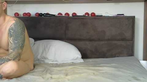 Video of a nude man with tattoos kneeling on a bed, head turned away, against a dark grey upholstered headboard. The room's minimalistic with a white wall and a shelf holding red objects.