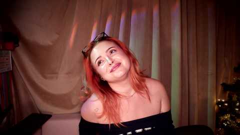 Media: Video of a smiling, light-skinned woman with long, vibrant red hair, wearing a black off-shoulder top. She stands against a backdrop of draped beige fabric, illuminated by colorful, abstract light reflections.