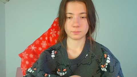 Video of a young woman with shoulder-length brown hair, wearing a dark green sweater with reindeer pattern, seated against a red and white snowflake-patterned backdrop.