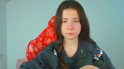 Video of a young girl with long, dark hair, wearing a grey sweater adorned with cartoon characters, sitting against a red blanket with snowflake patterns, against a light blue wall.
