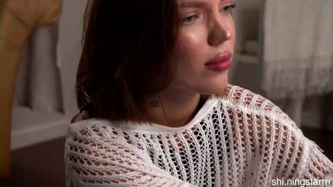 A video of a Caucasian woman with long, dark hair, wearing a white, knitted sweater, sitting in a softly lit room with a blurred background.