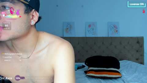 Video of a shirtless young man with glasses and a backward baseball cap in a modern bedroom with light-colored walls, a bed, and colorful art.
