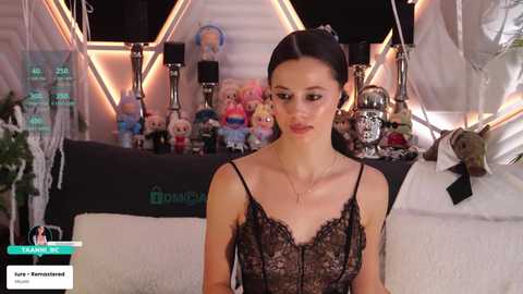 Video of a slender woman with fair skin and dark hair, wearing a black lace dress, standing amidst a colorful collection of plush toys in a dimly lit room.