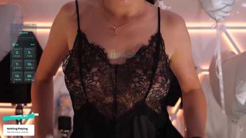 Video of a woman in a black lace camisole, revealing a tattoo on her chest, standing in a dimly lit room with blurred medical equipment and a digital display.