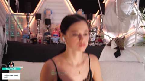 Video of a woman with fair skin and dark hair in a black lace dress, standing in a dimly lit room with a staircase, stuffed toys, and a white bed.