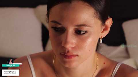 Video of a fair-skinned woman with dark hair pulled back, wearing a white camisole, sitting in a dimly lit room with white pillows, looking contemplative.