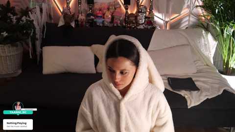 Video of a woman in a white bathrobe, sitting on a black sofa in a cozy, dimly lit living room with a fireplace and decorative candles.