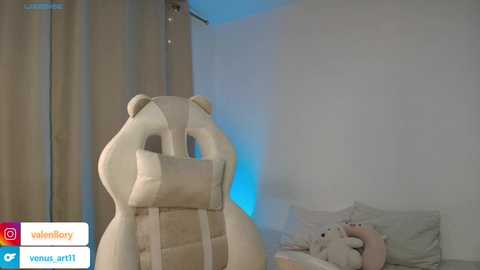 Video of a plush, white bear chair with a blue light, in a minimalist, white-walled bedroom with a plush teddy bear on a bed.