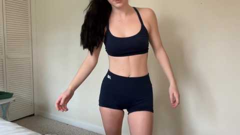 Video of a slender, fair-skinned woman with long black hair, wearing a black sports bra and high-waisted black shorts, standing in a beige-walled room with a closet and bed visible in the background.
