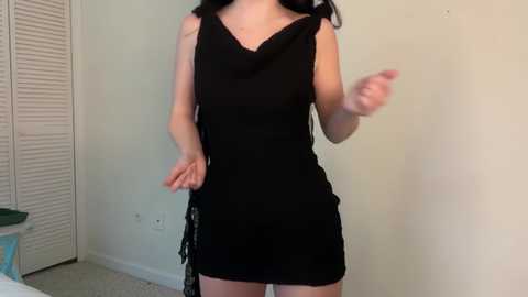 Video of a woman with long black hair wearing a black, sleeveless, V-neck dress. She stands in a room with white walls, a closed white louvered door, and a green object on a table in the background.