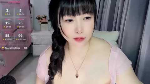 A video of an Asian woman with fair skin and long black hair, wearing a pink top and necklace, in a bedroom with a bed and curtains.