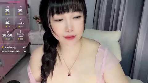 A young Asian woman with fair skin and long black hair in a braid, wearing a pink top, sits indoors. A live stream overlay shows viewers and chat.