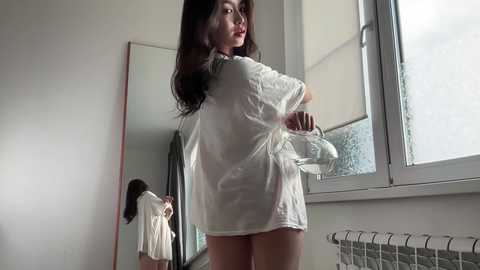 A video of a young woman with long dark hair, wearing a loose white shirt, standing near a large mirror in a dimly lit room with a window.