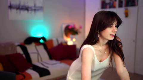 Video of a young woman with long brown hair, wearing a white top, in a modern living room with colorful pillows, abstract art, and a neon blue light.