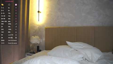 Video of a minimalist bedroom with a beige headboard, white pillows, and a vase of white flowers on a nightstand. A wall sconce emits warm light.