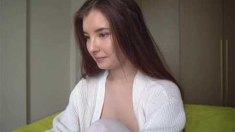 Media: Video of a young woman with long brown hair, fair skin, and light makeup, wearing a white knitted cardigan, sitting on a bed with a lime-green sheet. She gazes downward, appearing contemplative. Background features neutral-toned wardrobe.